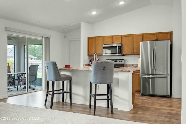 kitchen featuring a breakfast bar area, a kitchen island with sink, stainless steel appliances, wood finished floors, and brown cabinetry