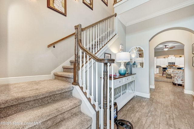 staircase featuring a ceiling fan, wood finished floors, arched walkways, crown molding, and baseboards