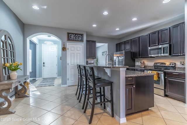 kitchen with arched walkways, a breakfast bar, appliances with stainless steel finishes, an island with sink, and dark stone countertops