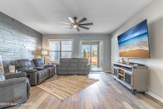 living area featuring light wood-style flooring, an accent wall, a ceiling fan, wooden walls, and baseboards