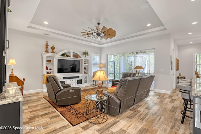 living area with a raised ceiling, a ceiling fan, light wood-type flooring, and baseboards