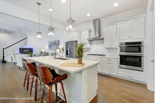 kitchen featuring wood finished floors, a sink, appliances with stainless steel finishes, crown molding, and wall chimney exhaust hood
