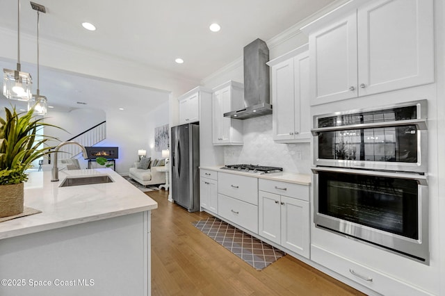 kitchen featuring light wood finished floors, wall chimney range hood, white cabinets, stainless steel appliances, and a sink
