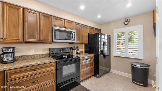 kitchen featuring brown cabinets, light tile patterned flooring, black appliances, light stone countertops, and baseboards