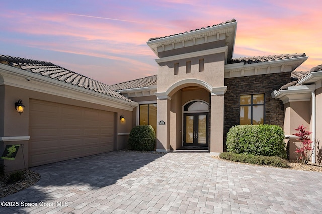 view of front of house featuring stone siding, an attached garage, decorative driveway, french doors, and stucco siding