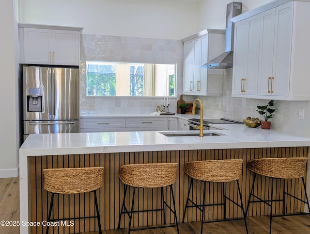 kitchen featuring a peninsula, decorative backsplash, white cabinetry, wall chimney range hood, and stainless steel fridge