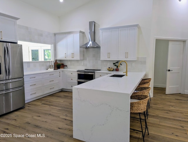 kitchen with a peninsula, a towering ceiling, stainless steel appliances, wall chimney exhaust hood, and a sink