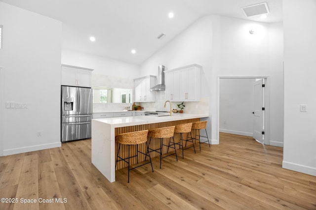 kitchen featuring high vaulted ceiling, stainless steel refrigerator with ice dispenser, a peninsula, wall chimney exhaust hood, and light countertops