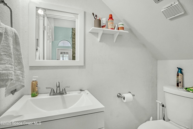 half bathroom with visible vents, toilet, lofted ceiling, vanity, and a textured wall