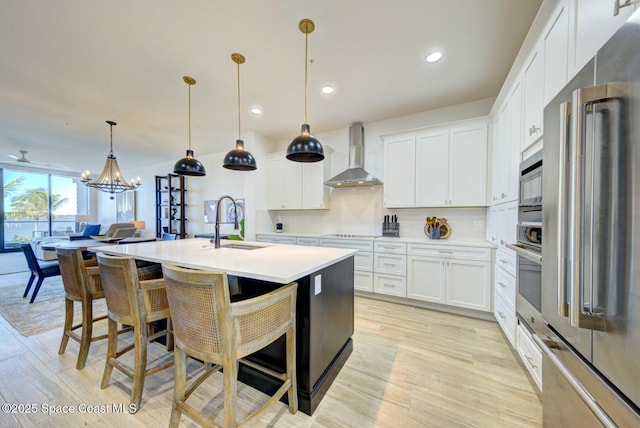 kitchen with a sink, stainless steel appliances, light countertops, wall chimney range hood, and light wood-type flooring