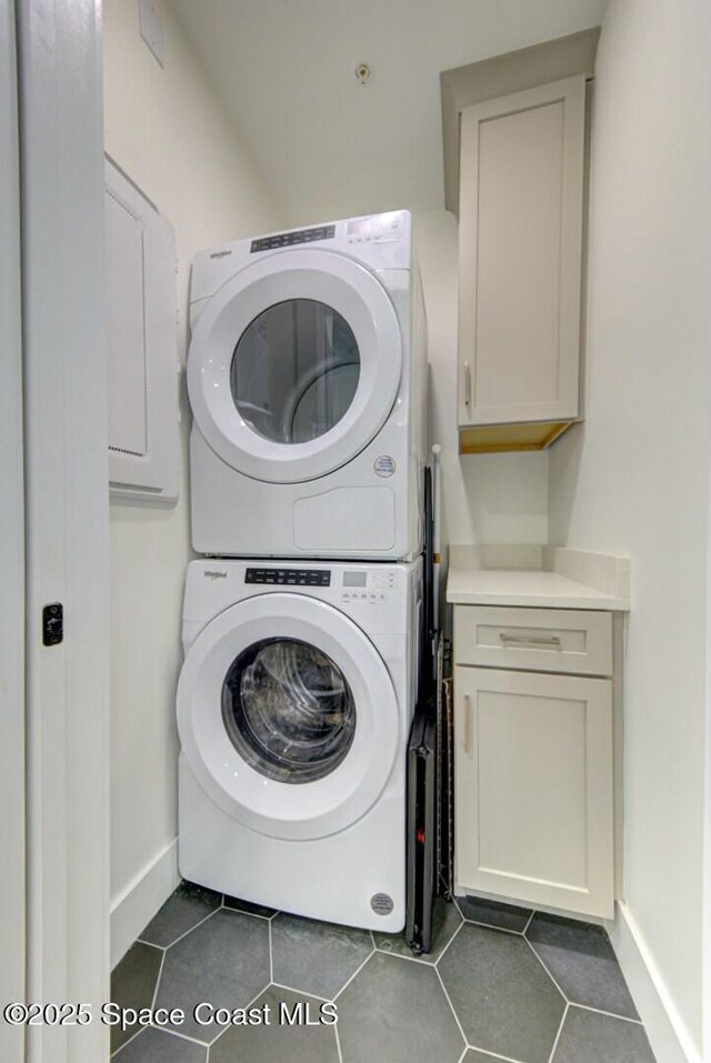 clothes washing area featuring dark tile patterned floors, cabinet space, stacked washer and clothes dryer, and baseboards