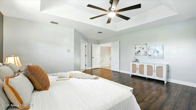 bedroom featuring visible vents, a ceiling fan, a tray ceiling, baseboards, and dark wood-style flooring