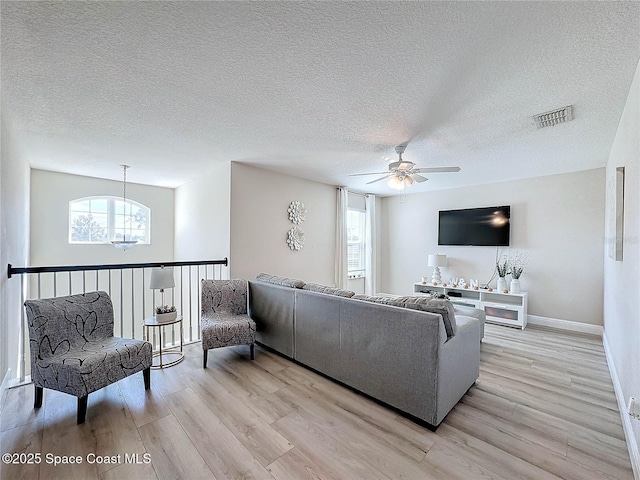 living room featuring visible vents, light wood-style flooring, ceiling fan with notable chandelier, a textured ceiling, and baseboards