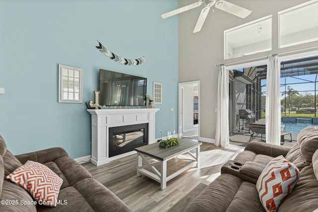 living room with wood finished floors, baseboards, a high ceiling, ceiling fan, and a glass covered fireplace