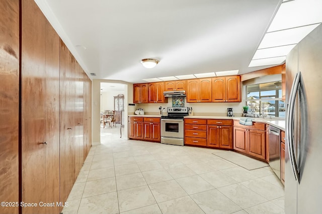 kitchen featuring light tile patterned flooring, stainless steel appliances, light countertops, under cabinet range hood, and brown cabinets