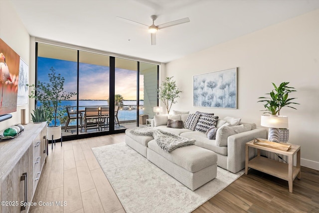 living area featuring baseboards, a water view, a wall of windows, light wood-style flooring, and a ceiling fan