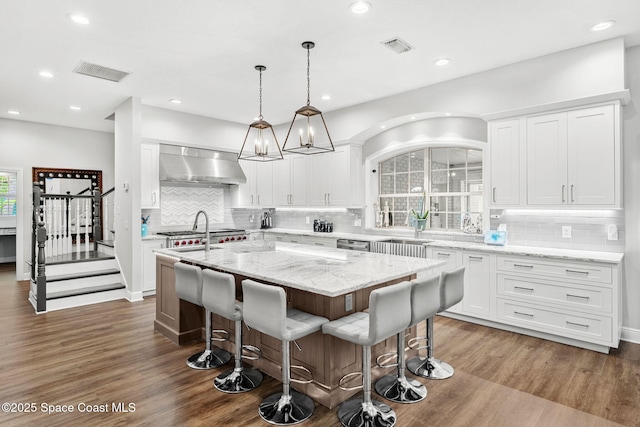 kitchen with wall chimney range hood, an island with sink, wood finished floors, and visible vents