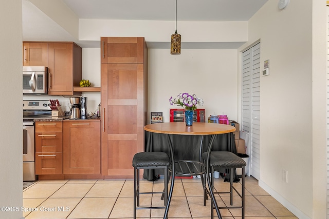 kitchen with dark countertops, brown cabinetry, light tile patterned floors, hanging light fixtures, and stainless steel appliances