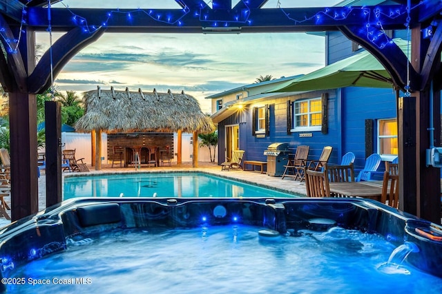 pool at dusk with area for grilling, an outdoor hot tub, fence, and outdoor dry bar