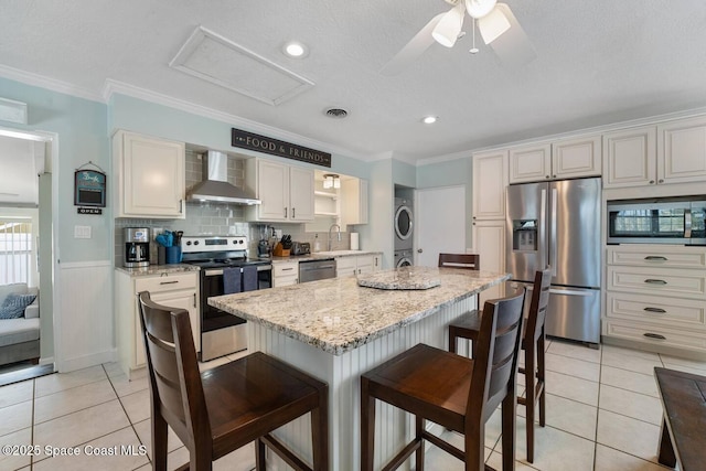 kitchen with ornamental molding, stacked washing maching and dryer, appliances with stainless steel finishes, a breakfast bar area, and wall chimney exhaust hood