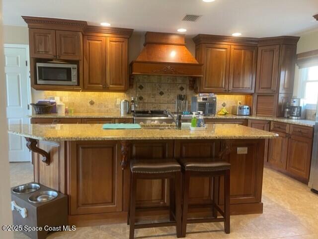 kitchen with light stone counters, a center island with sink, a breakfast bar, custom exhaust hood, and stainless steel microwave