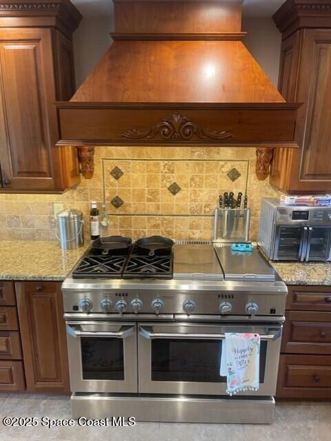 kitchen featuring decorative backsplash, range with two ovens, light stone counters, and custom range hood