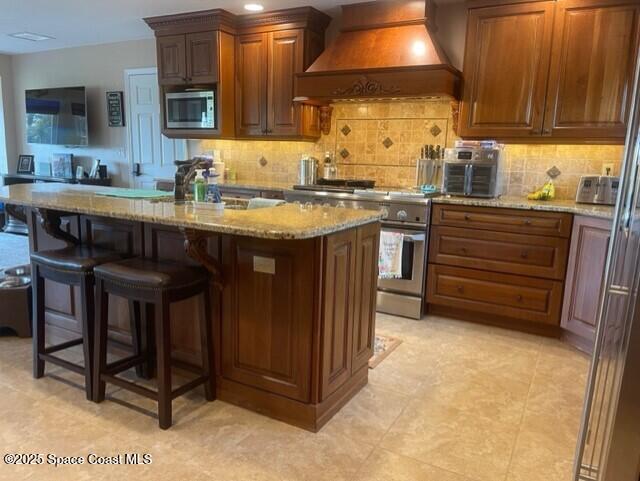 kitchen with light stone counters, a center island with sink, custom exhaust hood, appliances with stainless steel finishes, and tasteful backsplash