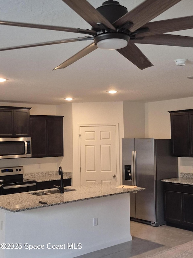 kitchen featuring a center island with sink, a sink, light stone counters, appliances with stainless steel finishes, and ceiling fan