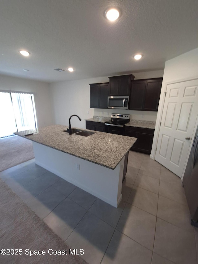 kitchen with a center island with sink, a sink, light stone counters, appliances with stainless steel finishes, and light tile patterned floors