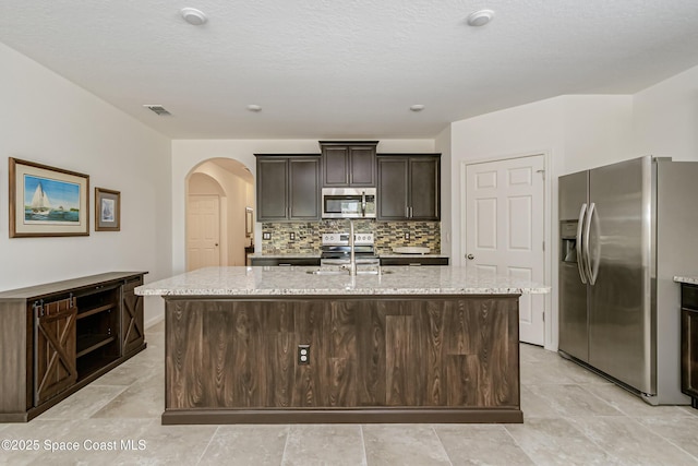 kitchen featuring a sink, backsplash, appliances with stainless steel finishes, arched walkways, and a kitchen island with sink