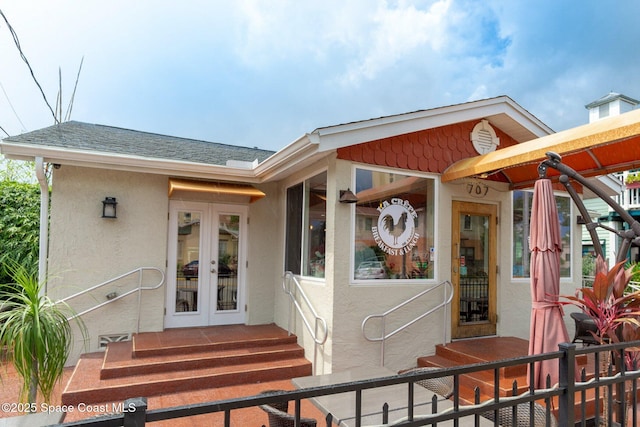 view of exterior entry featuring french doors, roof with shingles, and stucco siding