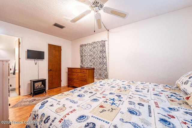 bedroom with visible vents, a ceiling fan, a textured ceiling, wood finished floors, and a wood stove