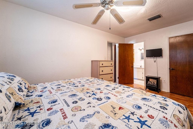 bedroom featuring visible vents, a textured ceiling, wood finished floors, ceiling fan, and a wood stove