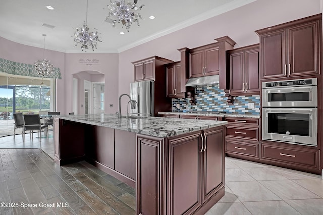 kitchen with arched walkways, decorative light fixtures, appliances with stainless steel finishes, under cabinet range hood, and backsplash