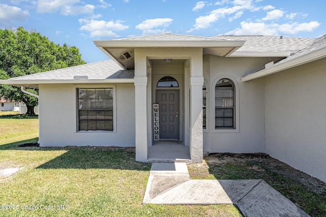 doorway to property featuring a shingled roof, a lawn, and stucco siding