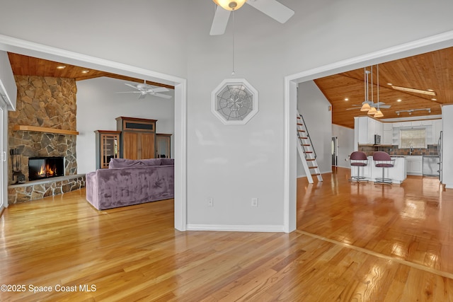 living room featuring a ceiling fan, light wood-type flooring, wooden ceiling, a stone fireplace, and stairs