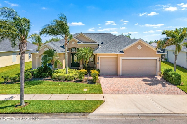 ranch-style house featuring decorative driveway, roof with shingles, a front yard, stucco siding, and a garage