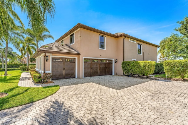 view of side of property with a tiled roof, an attached garage, a lawn, stucco siding, and decorative driveway