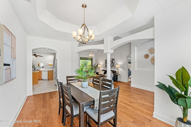 dining area featuring light wood finished floors, a raised ceiling, arched walkways, decorative columns, and a notable chandelier