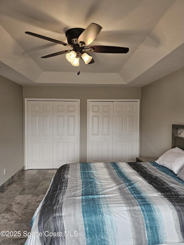 bedroom with a raised ceiling, ceiling fan, baseboards, and two closets