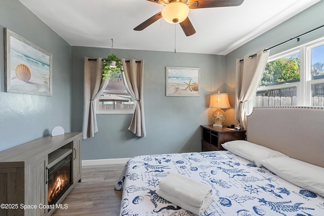 bedroom with wood finished floors, ceiling fan, baseboards, and a glass covered fireplace