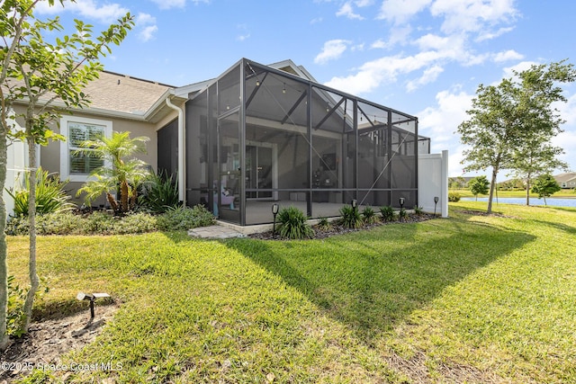 rear view of house with stucco siding, a shingled roof, a water view, glass enclosure, and a lawn