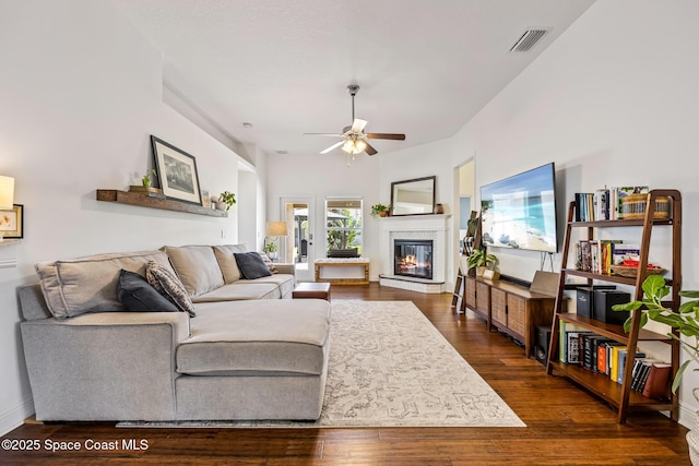 living area with dark wood-type flooring, a glass covered fireplace, visible vents, and ceiling fan