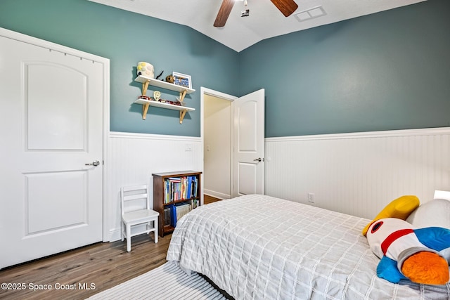 bedroom featuring a wainscoted wall, vaulted ceiling, wood finished floors, visible vents, and a ceiling fan
