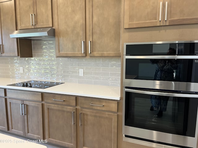 kitchen with under cabinet range hood, light countertops, black electric stovetop, stainless steel double oven, and backsplash