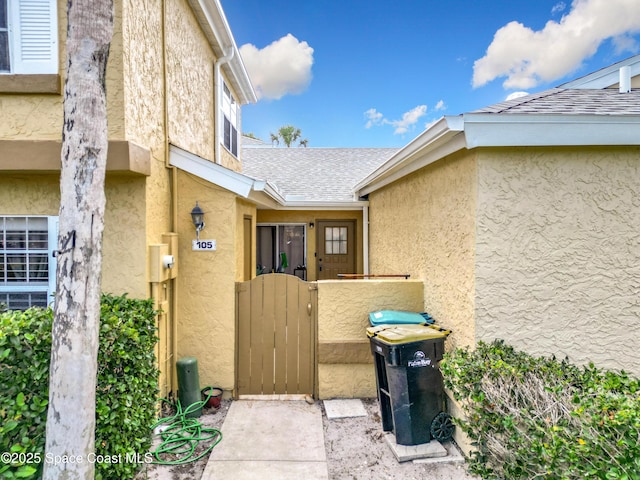 property entrance featuring a shingled roof, stucco siding, and a gate
