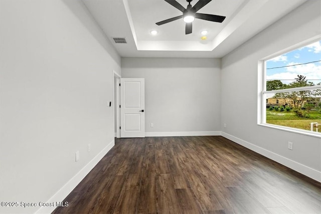 empty room featuring a tray ceiling, visible vents, a ceiling fan, baseboards, and dark wood-style flooring