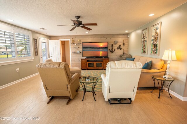 living room featuring wooden walls, a ceiling fan, light wood-style floors, baseboards, and a textured ceiling