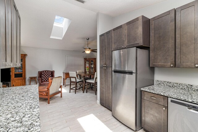 kitchen with stainless steel appliances, dark brown cabinets, a ceiling fan, light stone countertops, and vaulted ceiling with skylight