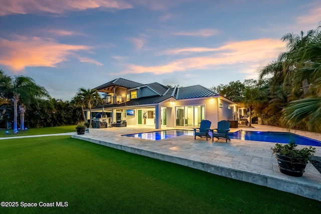 back of house at dusk with a balcony, a yard, a standing seam roof, and an outdoor pool